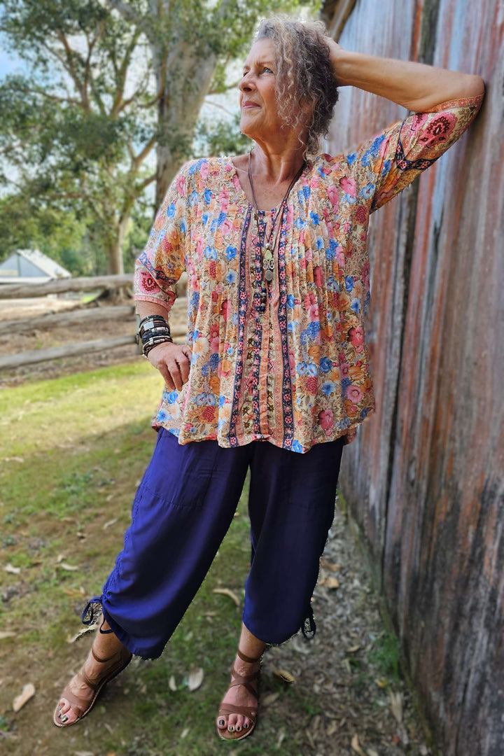 Woman in a colorful floral blouse and blue pants leaning against a wooden wall outdoors.