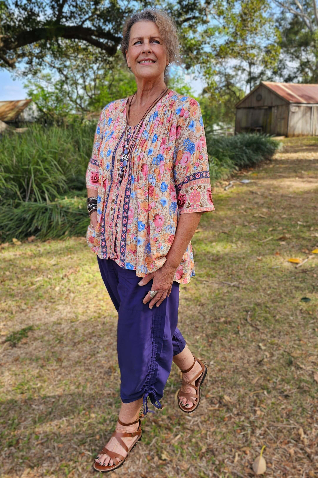 Woman in a colorful top and purple pants standing outdoors with trees and a building in the background.